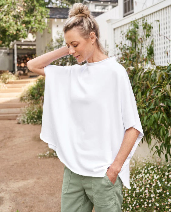 Displayed against a textured white background, the FRANK AND EILEEN - AUDREY FUNNEL NECK CAPELET IN WHITE exudes sophistication with its loose-fitting, short-sleeved design. Featuring a high, round neckline and minimalist style, this capelet is made from 100% Cotton Fleece by the renowned brand FRANK &amp; EILEEN, making it a refined addition to any wardrobe.