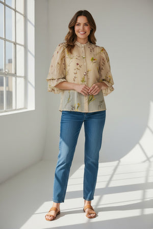 A woman stands in a sunlit room, smiling with her hands relaxed by her sides, wearing the MIRTO 1956 - BLOUSE WITH FLORAL PRINT from MIRTO 1956, paired with blue jeans and brown sandals.