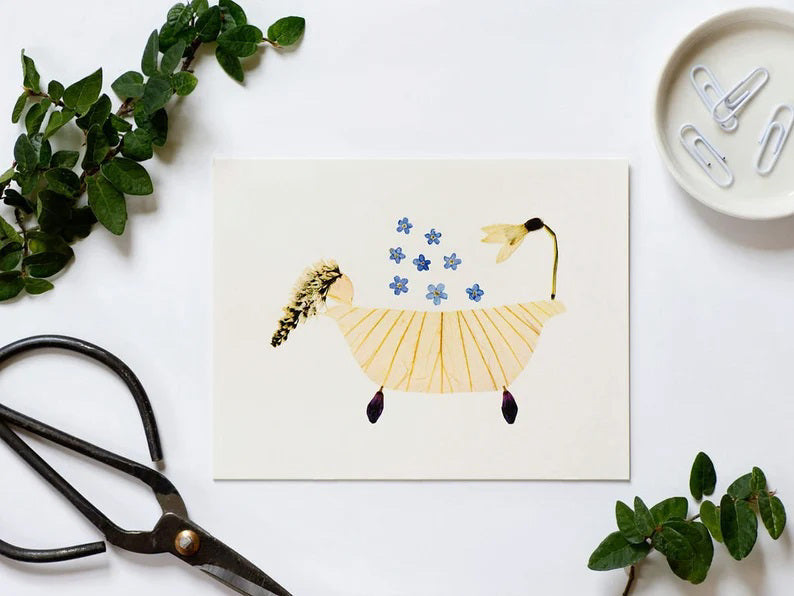 The PETAL PEOPLE PRESS - BUBBLE BATH CARD by PETAL PEOPLE PRESS showcases a whimsical horse crafted from yellow leaves with a petal body, white flower head, dark petal hooves, and feathery tail. Blue forget-me-not flowers are scattered above like falling rain in this pressed flower masterpiece.