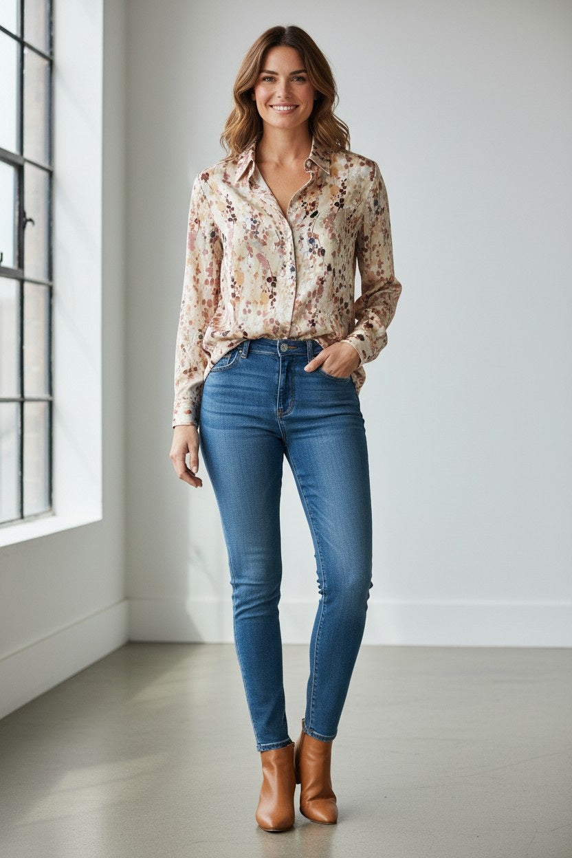A woman stands indoors by a window, smiling. She wears the DANI CONFEZIONI VLT’S BY VALENTINA Floral Viscose Blouse, paired with blue jeans and tan ankle boots. The room is bright with white walls and natural light.