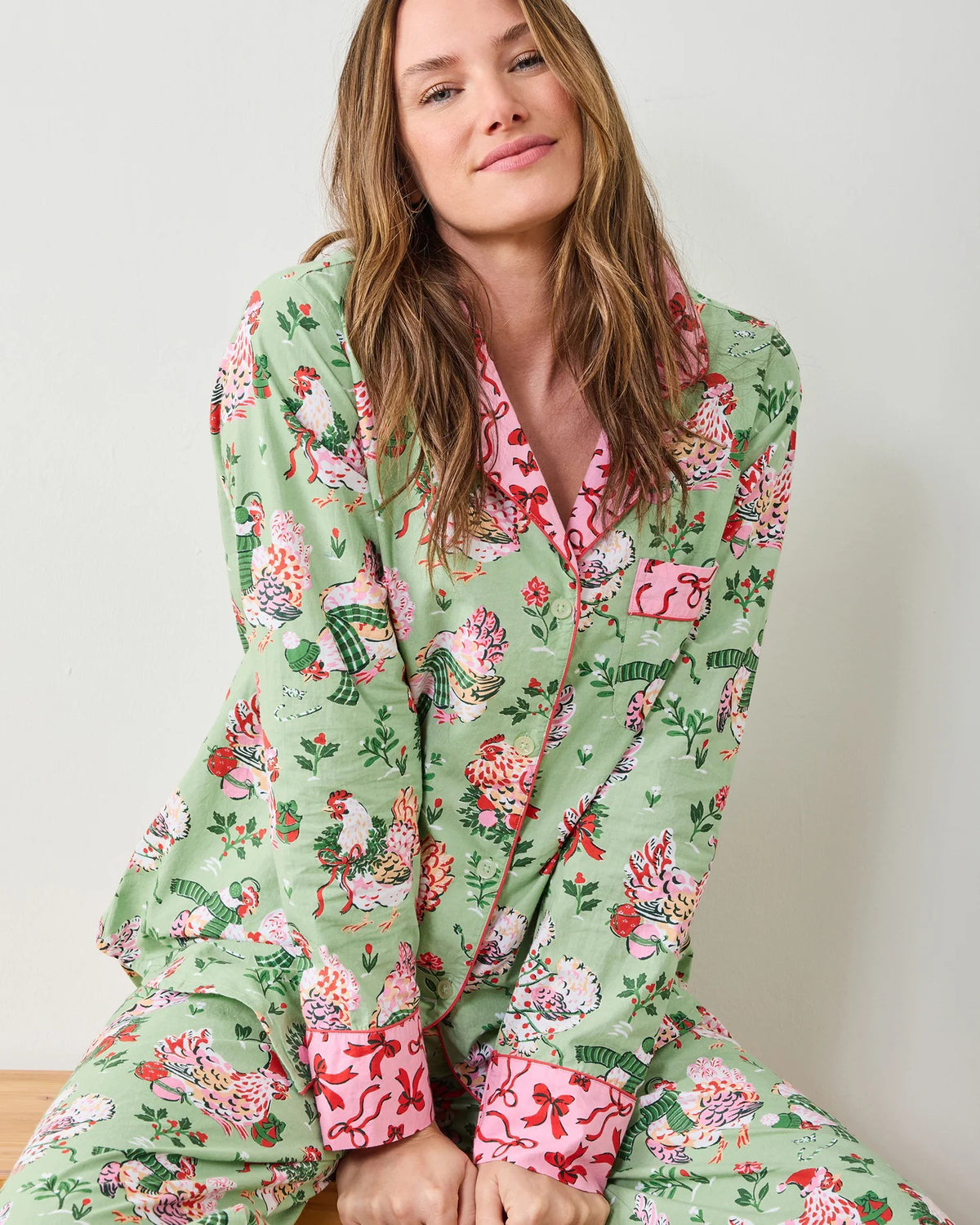 A woman stands barefoot against a white wall, smiling gently with her arms crossed. She wears the PRINTFRESH - HOLIDAY HENS LONG PAJAMA SET by PRINTFRESH, featuring light green organic cotton with a colorful floral and bird pattern and pink trim.