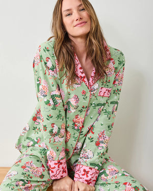 A woman stands barefoot against a white wall, smiling gently with her arms crossed. She wears the PRINTFRESH - HOLIDAY HENS LONG PAJAMA SET by PRINTFRESH, featuring light green organic cotton with a colorful floral and bird pattern and pink trim.