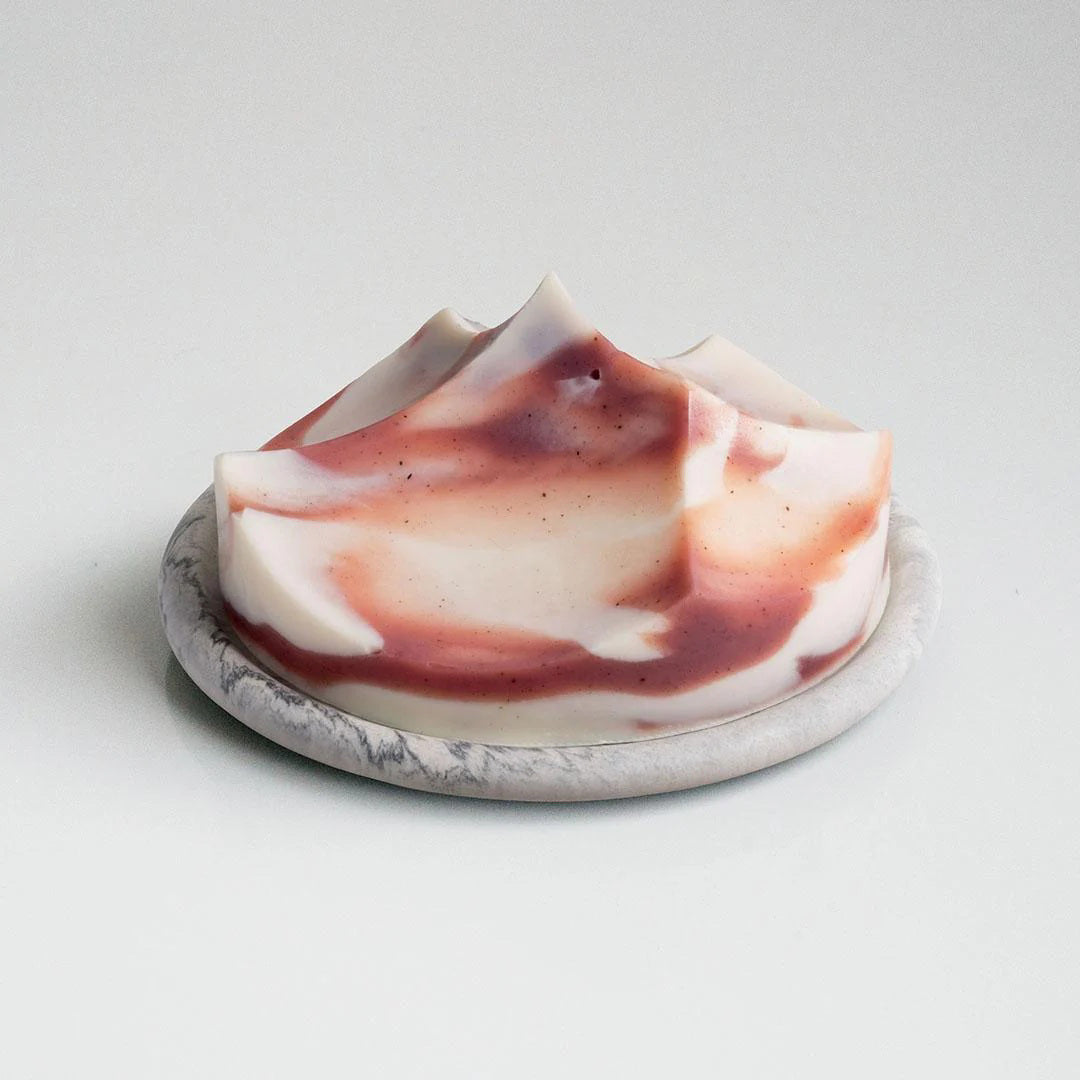 The UME STUDIO UME - LAVENDER CREME ERODE SOAP, with white and reddish-brown swirls and sculpted peaks, rests on a round marble-patterned soap dish against a plain background.