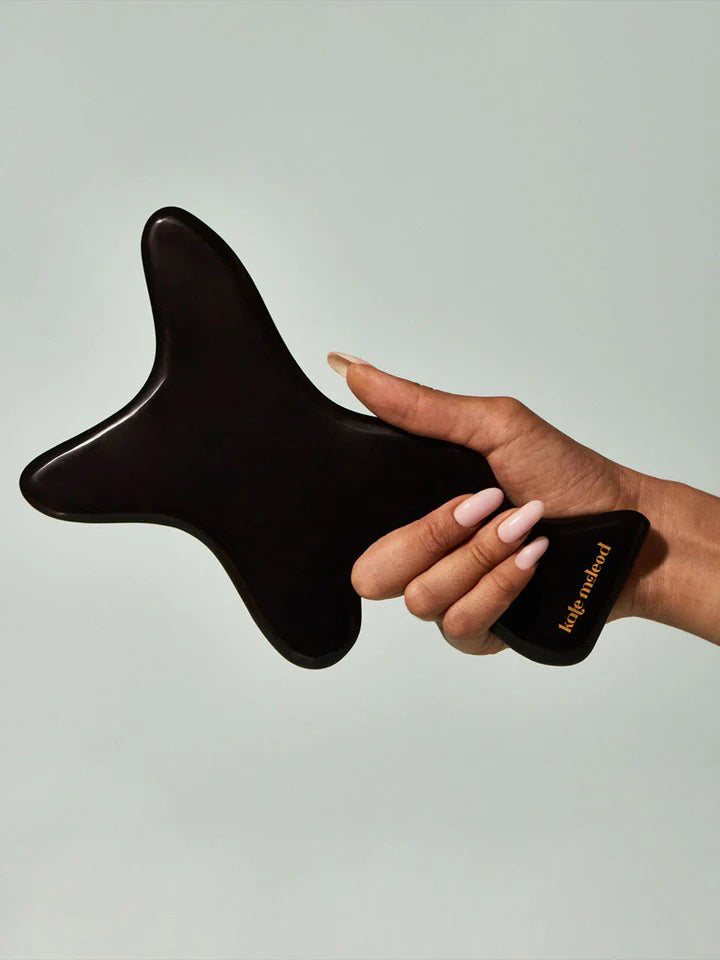 A woman with tied curly hair, wrapped in a beige towel, gently massages her forearm using the KATE MCLEOD - THE BODY GUA SHA, a black bian stone tool shaped like an oversized tuning fork. The neutral background exudes calm and relaxation.