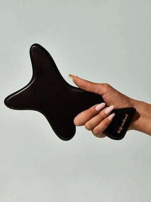 A woman with tied curly hair, wrapped in a beige towel, gently massages her forearm using the KATE MCLEOD - THE BODY GUA SHA, a black bian stone tool shaped like an oversized tuning fork. The neutral background exudes calm and relaxation.