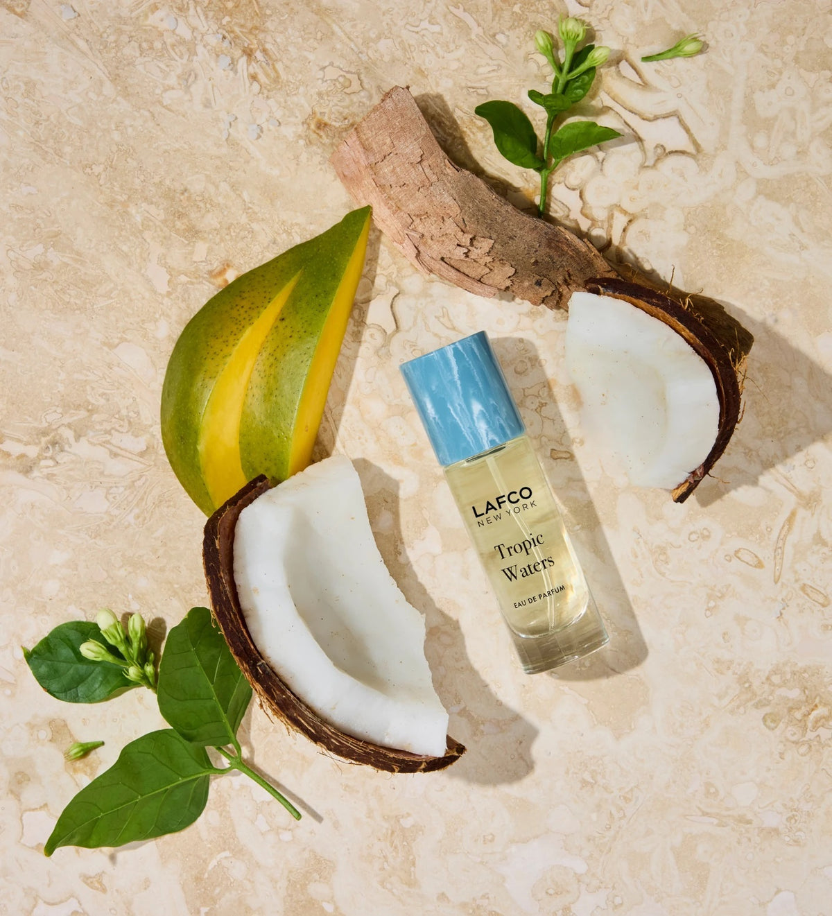 A LAFCO FINE FRAGRANCE, TROPIC WATERS 10ML box stands upright in a halved coconut, water splashing around it against a neutral background—an evocative display for this long-lasting scent inspired by the essence of coconut water.