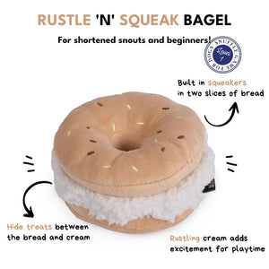 A small, fluffy white dog sniffs the CHEWFFON BAGEL PLUSH SQUEAKY DOG TOY, a large, round, beige plush toy resembling a sandwich, set against a plain light beige background.