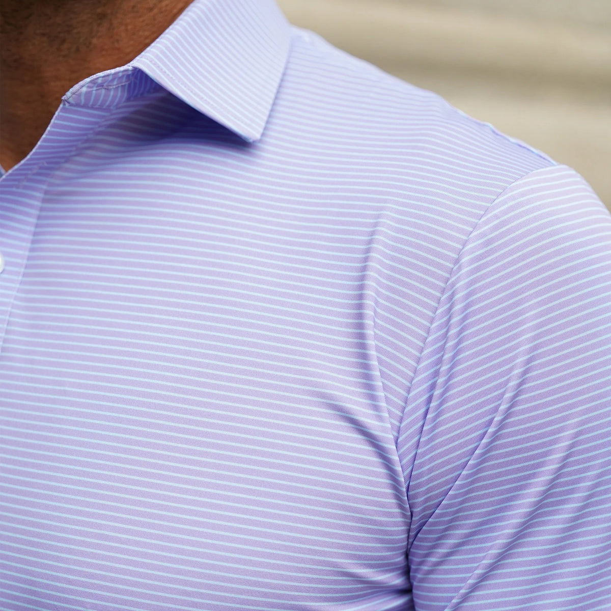 A man wearing sunglasses, a navy blazer, beige pants, and the COLLARS AND CO - Featherweight Flex Polo in Brookline Lavender with Turquoise Stripe stands outdoors in a leafy area, looking to the side and buttoning his jacket.