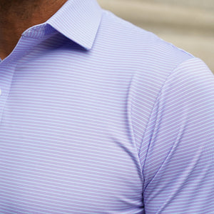 A man wearing sunglasses, a navy blazer, beige pants, and the COLLARS AND CO - Featherweight Flex Polo in Brookline Lavender with Turquoise Stripe stands outdoors in a leafy area, looking to the side and buttoning his jacket.