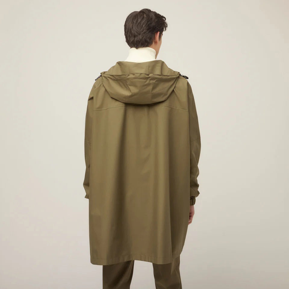 A person models the TILLEY - PACKABLE HOODED PONCHO in olive green, layered over a white turtleneck, standing against a plain light background and gazing to the side with a neutral expression. Brand: TILLEY.