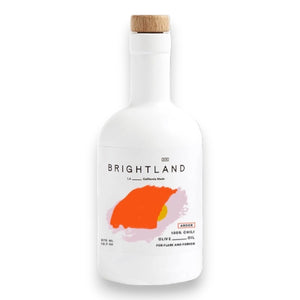 A white bottle with a cork lid, labeled "BRIGHTLAND" in black text. The label also features an orange-red abstract shape with the words "100% Chili Olive Oil" and notes "For flame and fervor." Made from Heirloom Frantoio Olives, this minimalist design stands against a plain background.