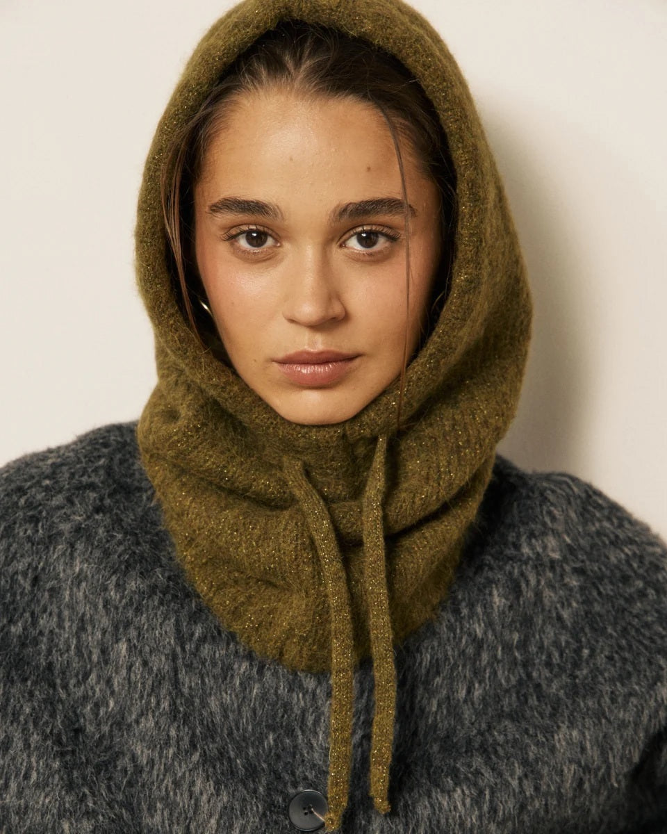 A woman wearing a fuzzy gray coat and the HARTFORD - MEVA HOOD IN KHAKI by HARTFORD—a cozy, merino wool balaclava crafted in Italy with drawstrings—looks directly at the camera against a plain background, adding both warmth and elegance to her winter style.