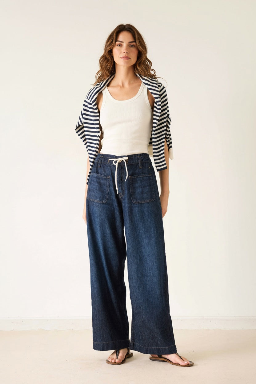 Wearing the MERSEA Londonberry Drawstring Jeans in Night Sky—a pair of wide-leg dark denim jeans with a comfortable drawstring waist—from MERSEA, styled with a cream sleeveless top, a striped shirt draped over the shoulders, and brown sandals, standing against a plain light background.