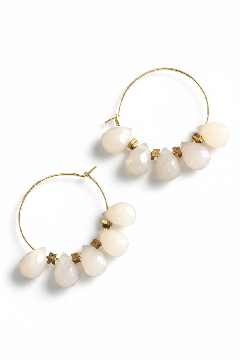 The DAVID AUBREY - DYED JADE MINI HOOP EARRINGS feature gold-plated hoops embellished with five teardrop-shaped, opaque white stones on each side, gracefully suspended from the hoops and displayed on a dark textured surface. Delicate gray jade beads from DAVID AUBREY accentuate the design, infusing these statement earrings with an elegant touch.