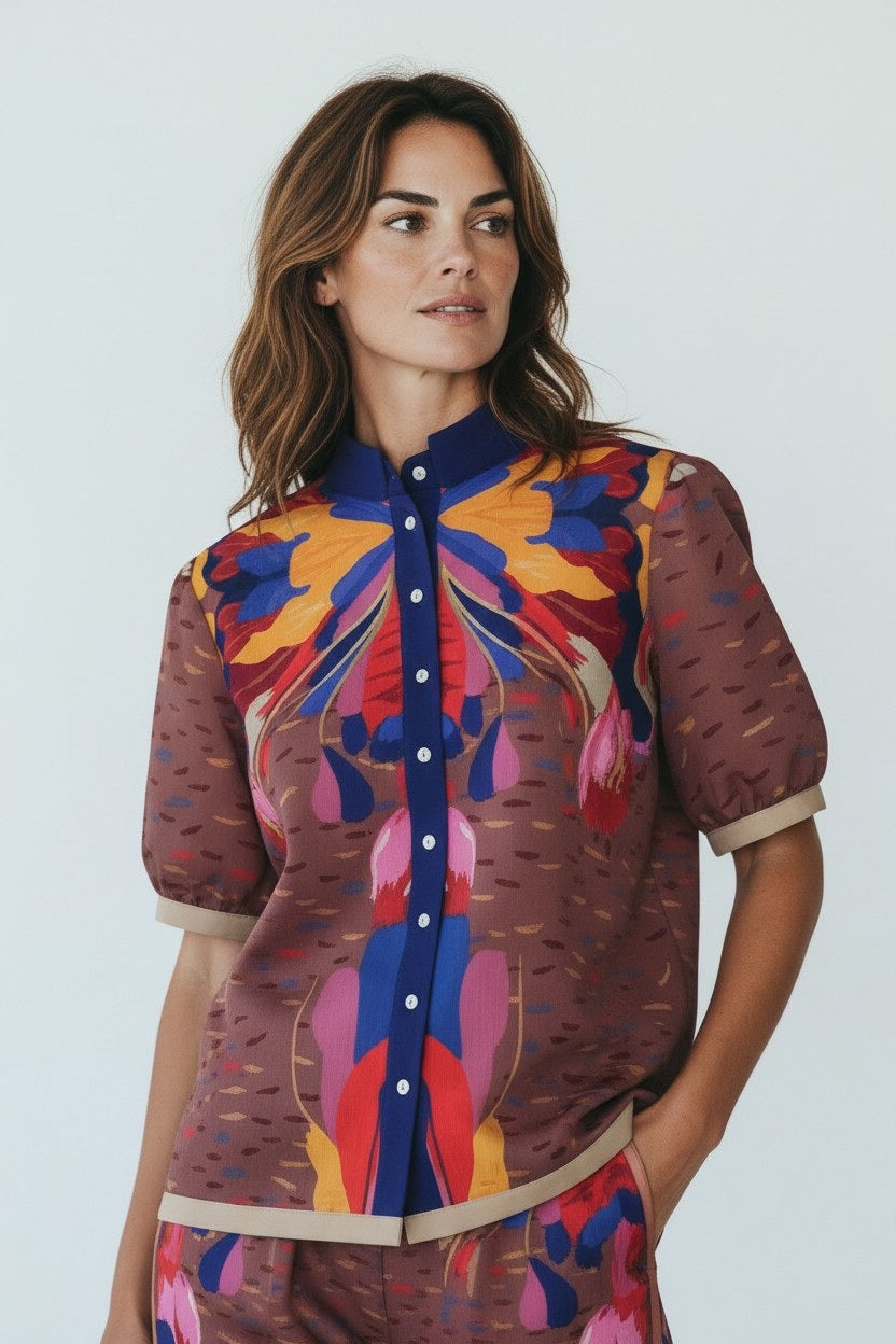 A woman stands indoors in natural light, wearing the FRANCO FERRARI Cotton Print Short Sleeve Blouse with matching loose pants. The set features a statement print in brown, blue, yellow, and pink. She completes her look with sandals.