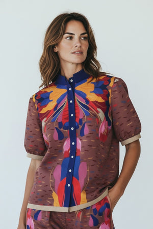 A woman stands indoors in natural light, wearing the FRANCO FERRARI Cotton Print Short Sleeve Blouse with matching loose pants. The set features a statement print in brown, blue, yellow, and pink. She completes her look with sandals.