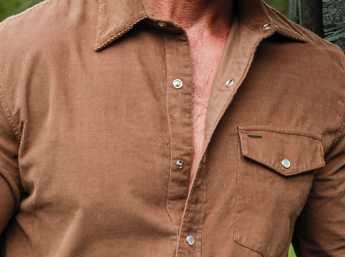 A man with short gray hair and a beard stands against a white background, wearing the CRIQUET Corduroy Pearl Snap Shirt in Walnut with rolled-up sleeves and dark jeans, smiling slightly with one hand in his pocket.