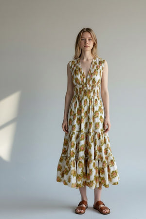 A woman stands against a plain light background wearing the HOPE FOR FLOWERS Tiered Peasant Maxi Dress in Pink Warped Ikat, a sleeveless, ankle-length dress with a shirred bodice and tiered skirt. She completes the look with brown sandals.