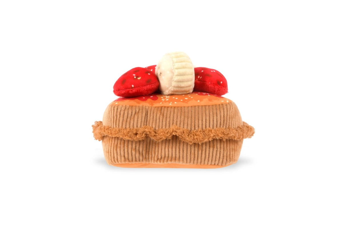 Close-up of the PLAY PET LIFESTYLE THICK ‘N FLUFFY FRENCH TOAST DOG TOY on a brown fabric with red and white patterns, pictured with plush strawberry and muffin toys.
