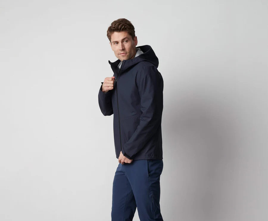 A man stands in front of a white wall wearing the MACK WELDON STORMCHASER JACKET with adjustable hood, paired with a gray shirt, blue pants, and black sneakers. He looks to the side while holding the jacket's zipper.