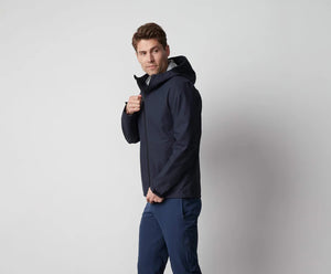 A man stands in front of a white wall wearing the MACK WELDON STORMCHASER JACKET with adjustable hood, paired with a gray shirt, blue pants, and black sneakers. He looks to the side while holding the jacket's zipper.