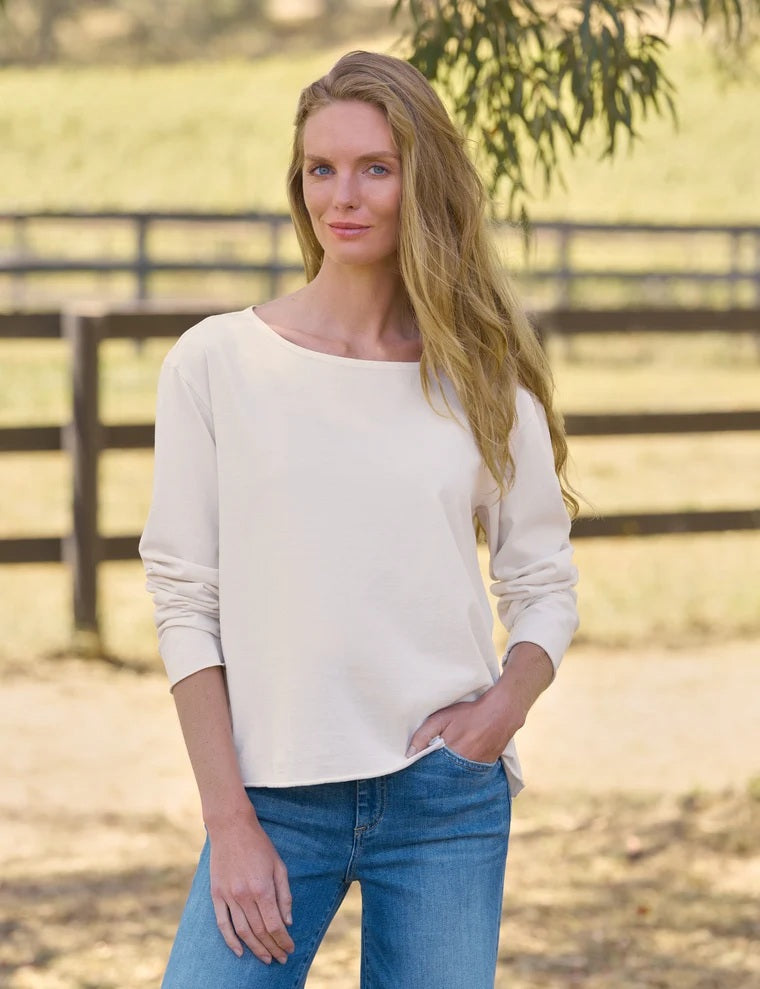 A person with long blonde hair, dressed in the FRANK & EILEEN ELLA Long Sleeve Boatneck Tee in Vintage White from the California Heritage Collection and blue jeans, stands outdoors on a sunny day, glancing over their shoulder. A wooden fence and trees provide a scenic background.