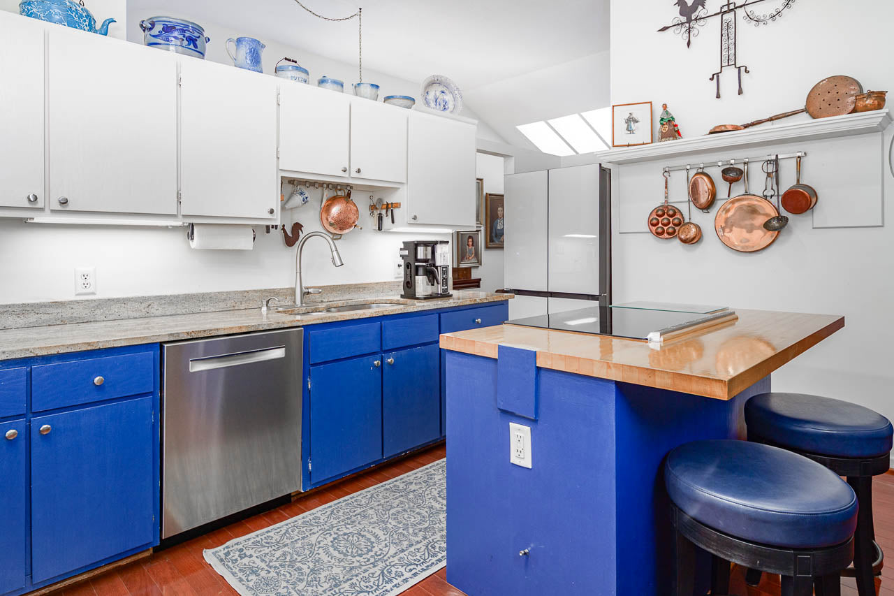 A kitchen with blue lower cabinets, white upper cabinets, stainless steel appliances, a wooden countertop island with two blue stools, and copper pots hanging on the wall. Blue decorative items are on top of the cabinets.
