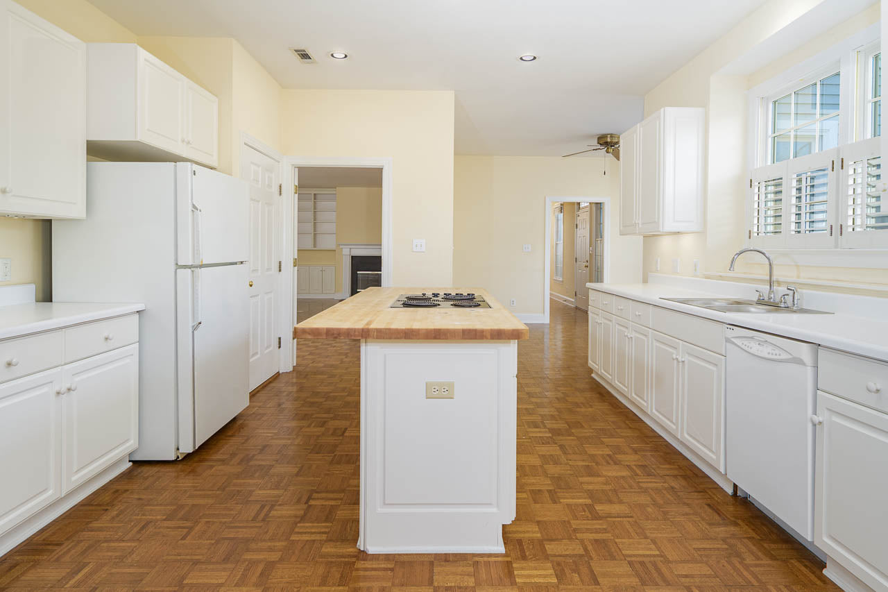Bright kitchen with white cabinets and appliances, a central island with a built-in cooktop, parquet wood flooring, and large windows allowing natural light into the space.