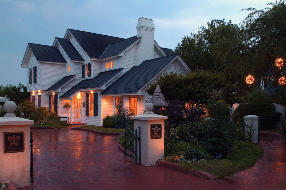 A large white two-story house with warm lights glowing from the windows, surrounded by lush gardens and trees, sits on a brick driveway at dusk. A sign and gate are visible at the entrance.