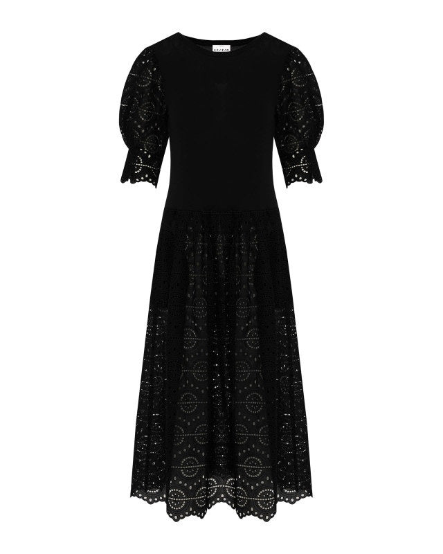 A person stands against a plain background wearing the SFIZIO - BRODERIE ANGLAISE MIDI DRESS by SFIZIO, which features elbow-length puffed sleeves and a midi-length skirt adorned with intricate lace patterns. They are also wearing black ankle-strap sandals.