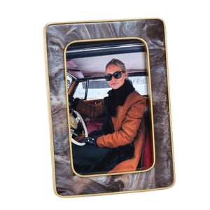 A person wearing sunglasses and a brown coat sits in a vintage car, holding the steering wheel. The photo is displayed in the ZODAX Marbled Hazel Resin Frame with Antique Brass Trim (4x6), featuring gold accents.