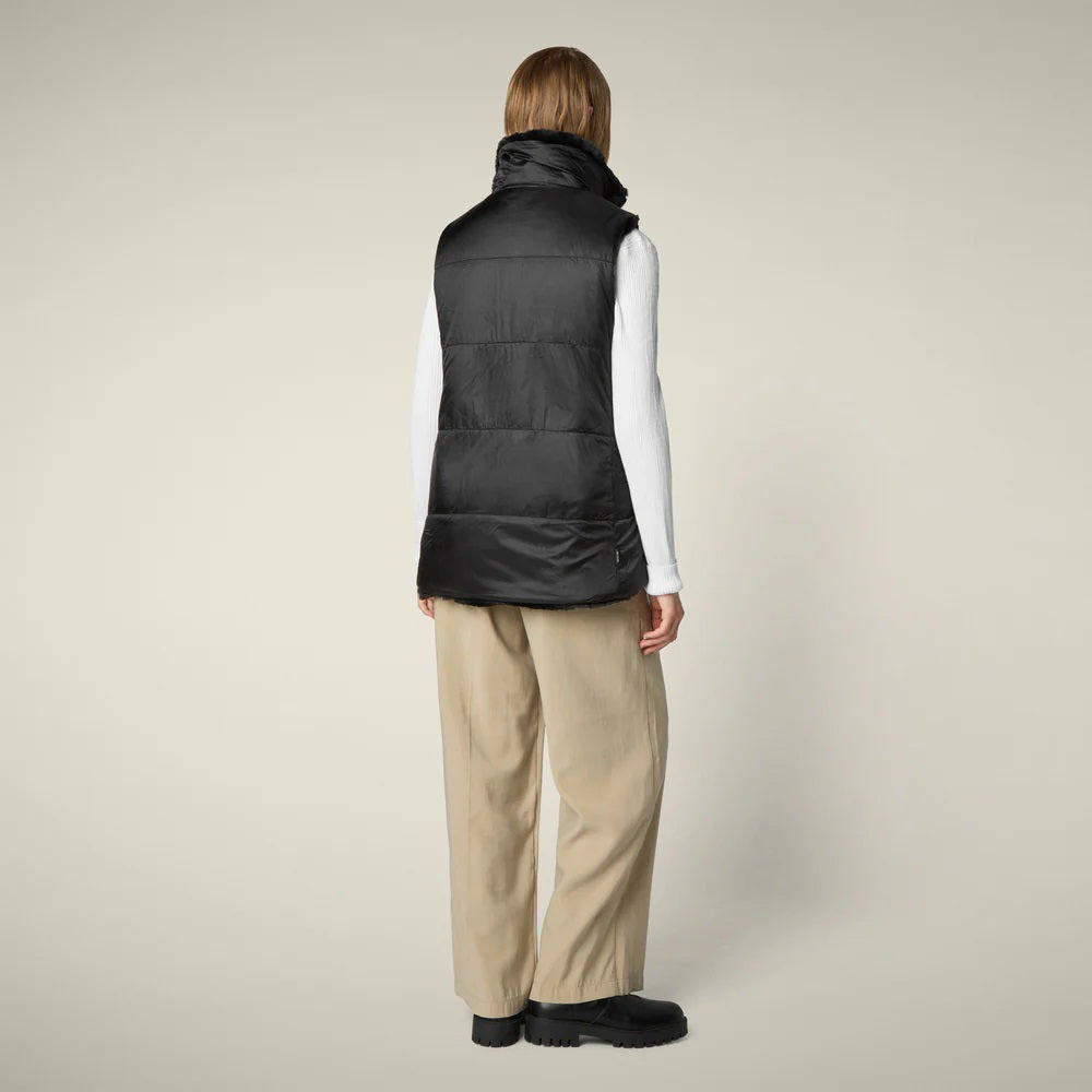 A woman with straight blonde hair wears the SAVE THE DUCK NIVES REVERSIBLE VEST over a white long-sleeve top, beige wide-leg pants, and black shoes, standing against a plain background and highlighting the vest&#39;s chic style.