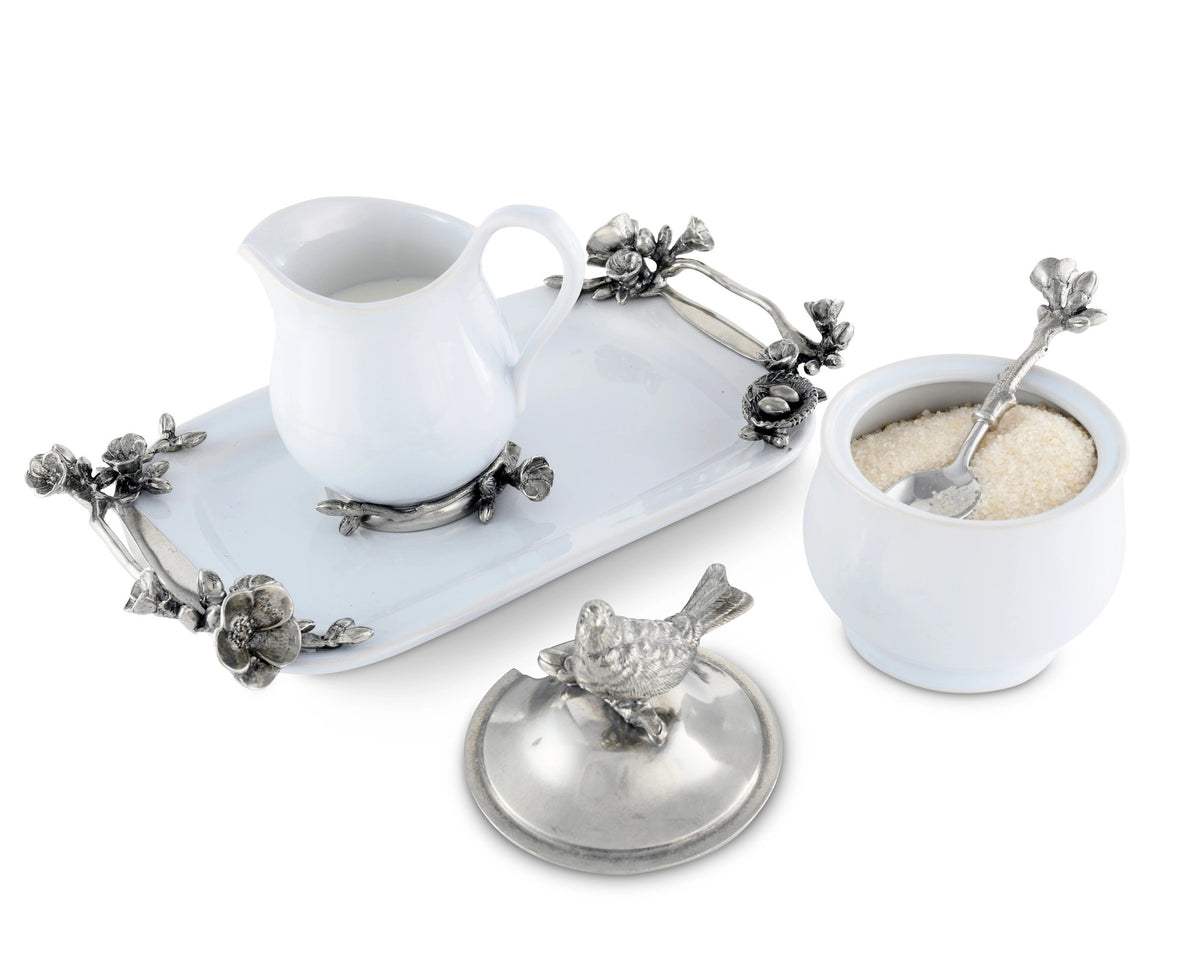The SONG BIRD CREAMER SET by VAGABOND HOUSE features a white ceramic sugar bowl with a lid, adorned with a bird figurine on top, paired with a matching cream pitcher. Both items rest on a decorative tray with metallic floral embellishments and include a small spoon with an ornate handle.