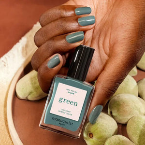A hand with short, oval-shaped nails painted in MANUCURIST INC’s sophisticated GREEN™ Nail Polish in Sage—a chic gray vegan shade—is held in front of bright pink-red flower petals.