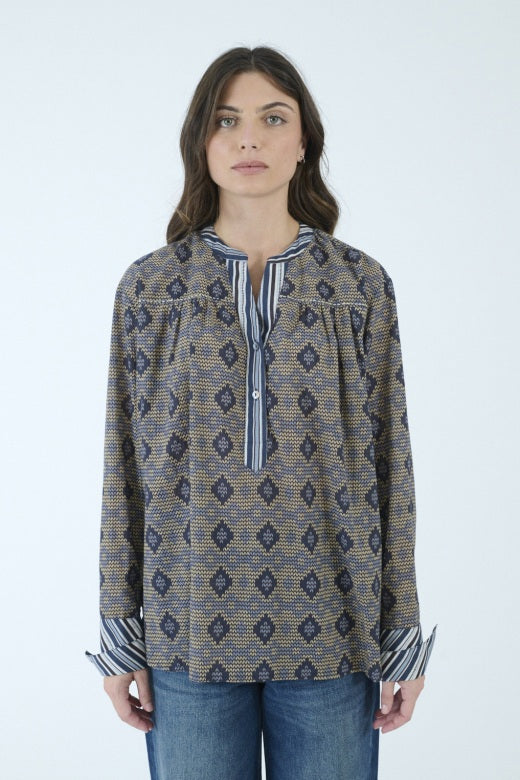 A person with long brown hair, seen from behind, wears the STORIATIPIC MARGOT BLOUSE IN BLUE by STORIATIPIC—a patterned cotton shirt featuring a mix of vertical stripes and diamond shapes in blue and beige tones—paired with blue jeans. The blouse is suitable for a gentle machine wash at 30 degrees.