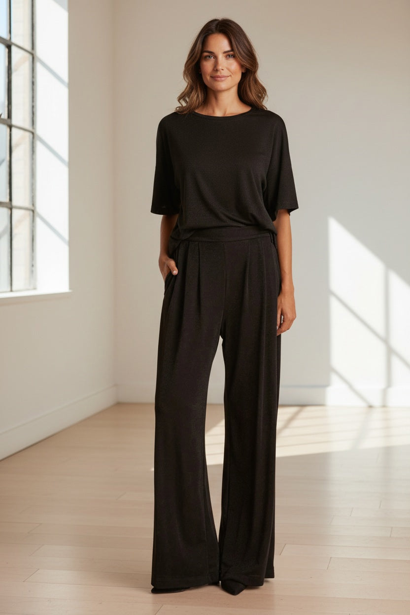 A woman stands against a neutral background wearing MOMONI Lanzarote Pant in Lurex Jersey, a loose black top, black shoes, and a black-and-white patterned scarf, with her hands in her pockets.