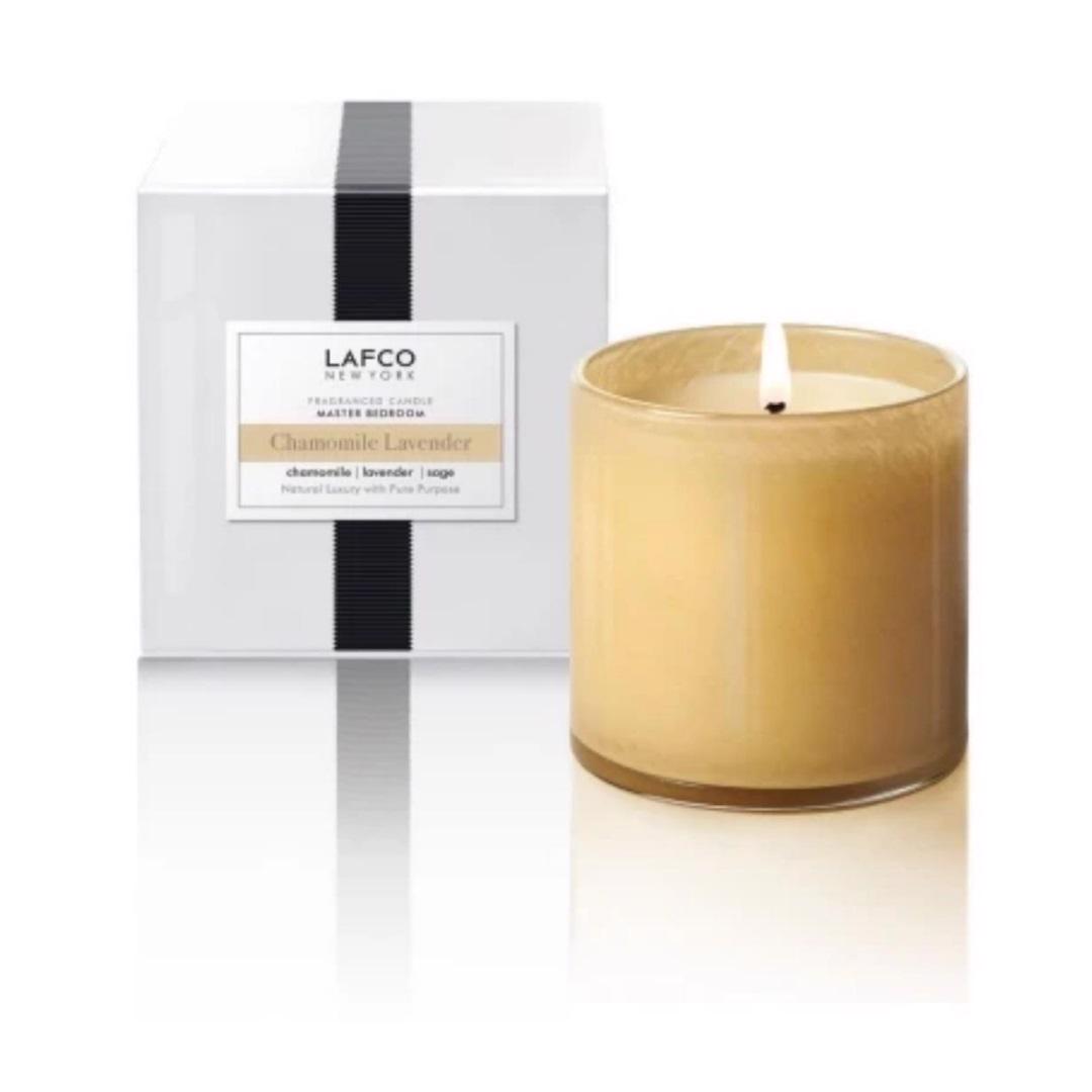 A burning LAFCO - Chamomile Lavender, Bedroom candle sits beside its elegant white box adorned with a black ribbon. The label on the box details the scent and notes. Hand-poured, this luxurious LAFCO candle boasts an exquisite lavender fragrance in a simple, sleek glass jar.
