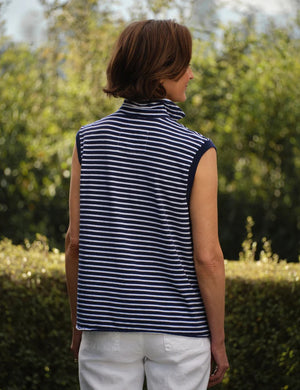 A woman with short brown hair stands outdoors against a backdrop of blurred greenery, wearing the FRANK & EILEEN OLIVIA Popover Henley Tank in Vintage Jersey Yacht Stripe—a sleeveless navy and white striped shirt with a popped collar.