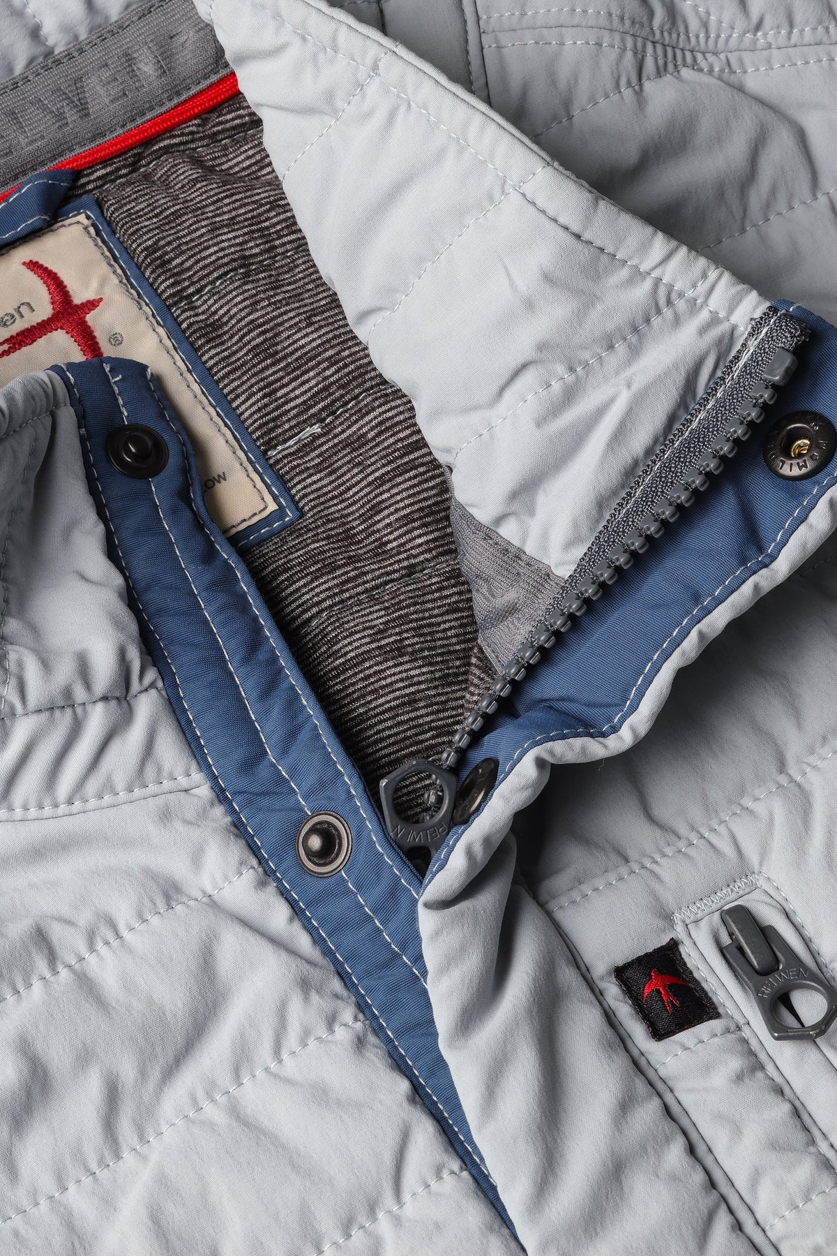 A person models the RELWEN - WINDZIP VEST IN GHOST GRAY over a gray sweater, plaid shirt, and dark blue jeans, standing with one hand in their pocket against a white background for extra layered warmth.