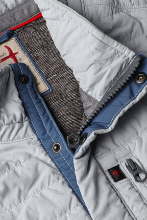 A person models the RELWEN - WINDZIP VEST IN GHOST GRAY over a gray sweater, plaid shirt, and dark blue jeans, standing with one hand in their pocket against a white background for extra layered warmth.