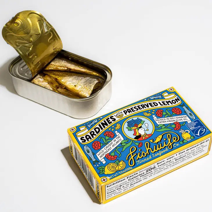 A vibrant, rectangular tin of FISHWIFE - SARDINES &amp; PRESERVED LEMON from the Fishwife brand. The label showcases lively illustrations of sardines, a lemon, and decorative elements. It highlights that the sustainably wild-caught sardines are sourced from the Northeast Atlantic and packed in Spanish extra virgin olive oil. Net weight: 4.2 oz.