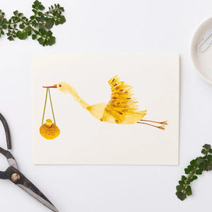 The PETAL PEOPLE PRESS - STORK CARD by PETAL PEOPLE PRESS features a stork crafted from yellow flower petals carrying a bundle, displayed on a pale wooden surface with an envelope peeking out behind it.