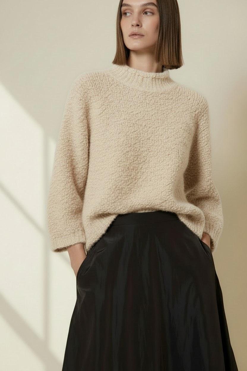 A woman stands indoors by a window, wearing the VICARIO CINQUE - MADEIRA Boucle Sweater in Light Taupe from VICARIO CINQUE, paired with a long black skirt and sneakers. Sunlight casts grid-like shadows on the wall.