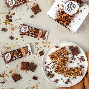 An 88 ACRES - DOUBLE DARK CHOCOLATE SEED + OAT BAR rests on a light wooden surface. The packaging is brown and white, showcasing images of dark chocolate chunks and oats. The label proudly highlights that it is gluten-free, vegan, and nut-free.
