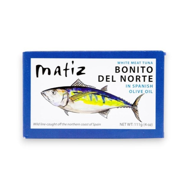 An opened can of MATIZ ESPANA - BONITO DEL NORTE IN SPANISH OLIVE OIL, in a blue box, is displayed with slices of baguette, lemon wedges, and a fork on a white surface.