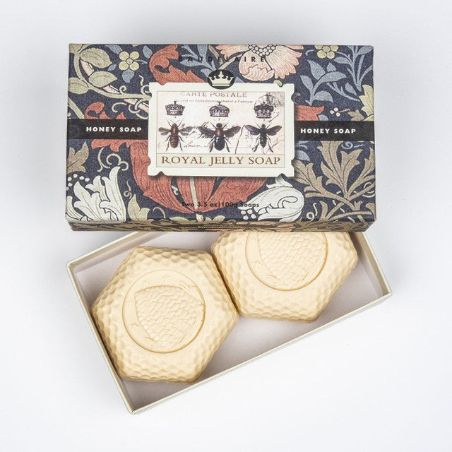 A box of BAUDELAIRE - ROYAL JELLY HONEY SOAP 3.5 2 BAR GIFT BOX is open to reveal two hexagonal bars embossed with a beehive pattern. The vintage botanical design on the box features bees and flowers, labeled as &quot;Honey Soap.&quot; Enhance hydration and achieve a glowing complexion with each use.