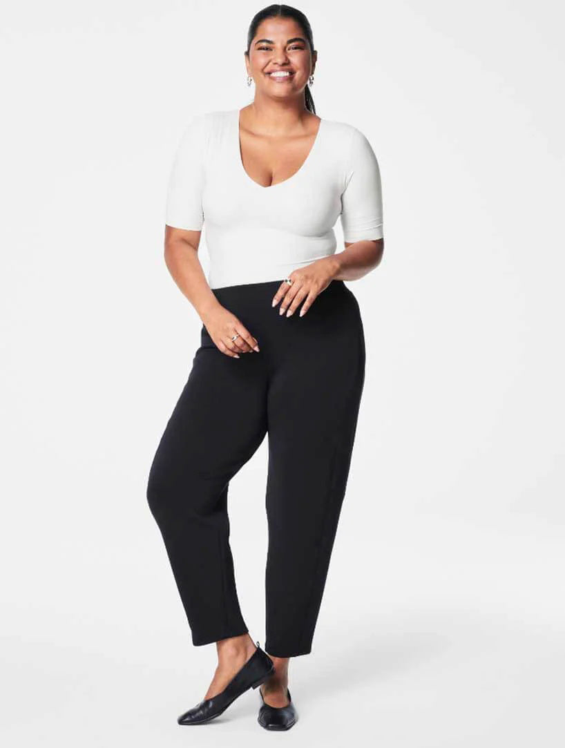 A woman with a cheerful smile stands in front of a white background. She is wearing a white, short-sleeve, V-neck top and SPANX - AireEssentials Barrel Pant in black made from modal and elastane. She completes the look with black flats and has her hands lightly clasped in front of her.