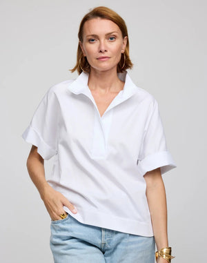 A woman with short light brown hair is seen standing against a plain light background, wearing the HINSON WU - CARA Elbow Sleeve Shirt in Stretch Polished Cotton White by HINSON WU. The oversized, loose-fit shirt features a polished cotton fabric with a collared neckline and elbow-length sleeves, and is paired with light blue jeans, large hoop earrings, and a gold bracelet. The machine-washable shirt offers effortless care.