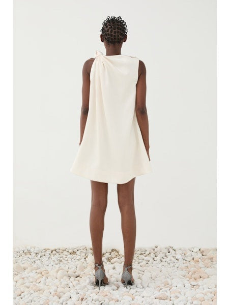 A person stands on white pebbles in the ELEH ONE SHOULDER KNOT DRESS by ELEH, an ivory minimalist dress with a draped shoulder detail and strappy heeled sandals, set against a plain white background.