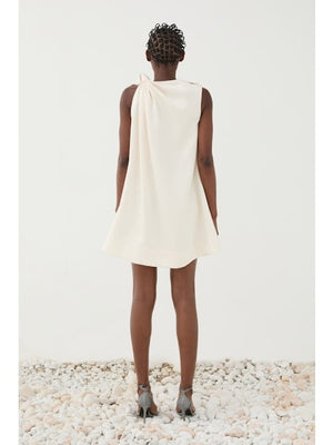 A person stands on white pebbles in the ELEH ONE SHOULDER KNOT DRESS by ELEH, an ivory minimalist dress with a draped shoulder detail and strappy heeled sandals, set against a plain white background.